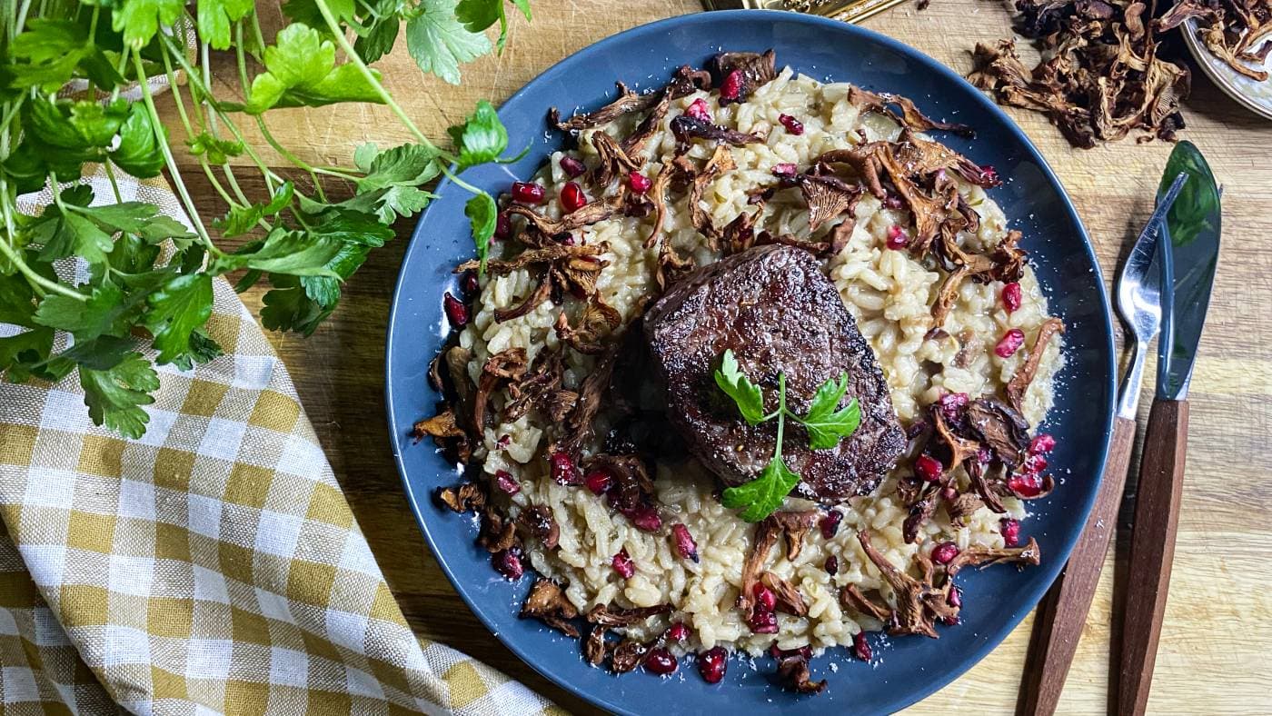 Ein hjortebiff på kantarellrisotto, pynta med steikte kantarellar, granateplekjernar og persille på ein blå tallerken.