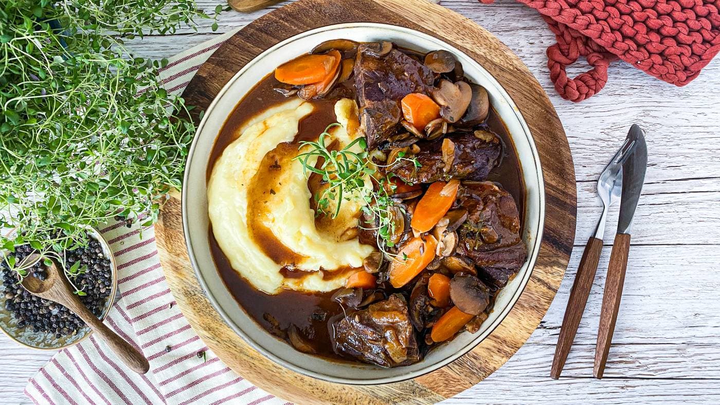 Hjortestek i mørk guinnessaus med gulrot og sopp, servert med potetmos i ei djup skål.